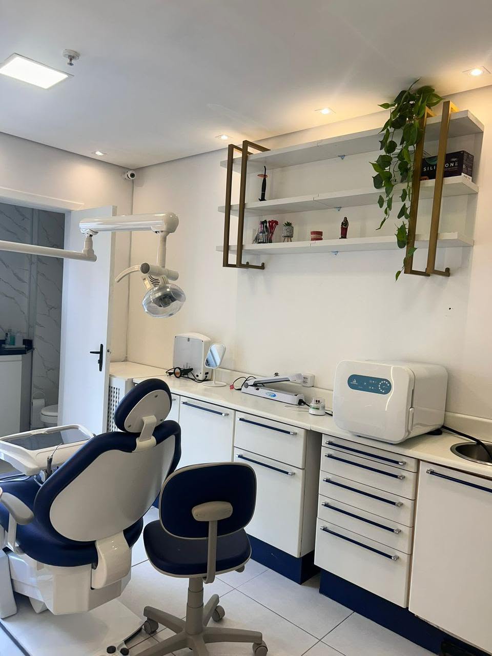 Sala de atendimento com cadeira odontológica azul, bancada e prateleiras.